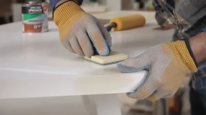 A man's hands in work gloves are shown in close-up, carefully sanding the edge of a white laminated furniture board with a sanding block.